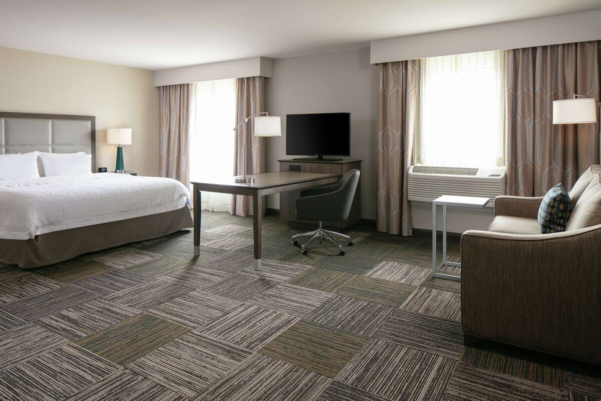 A hotel room with a bed, desk, chair, TV, and air conditioning unit.