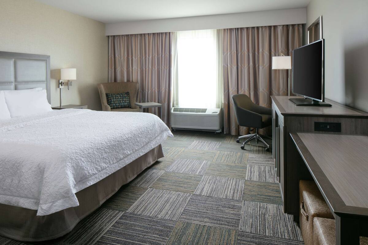 A hotel room with a large bed, a desk with a flat-screen TV, a chair, a small table, and an air conditioning unit.