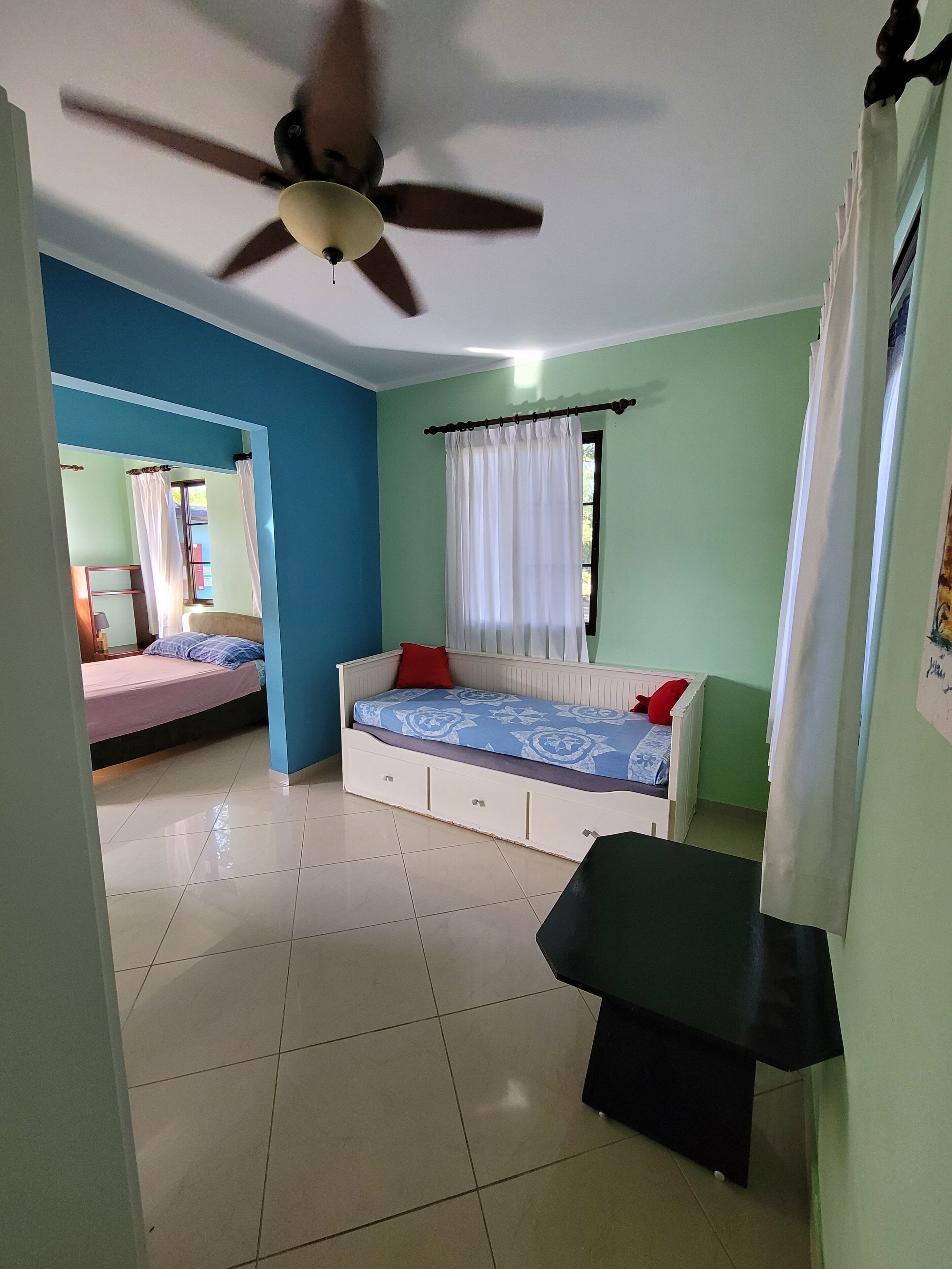 A room with a bed, a desk, a ceiling fan, and a window with curtains.