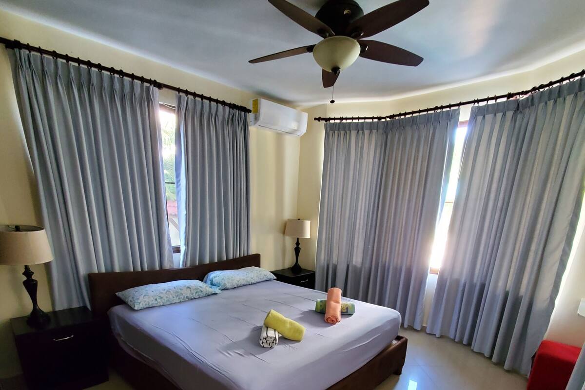 A hotel room with a bed, curtains, a ceiling fan, a nightstand, and a lamp.