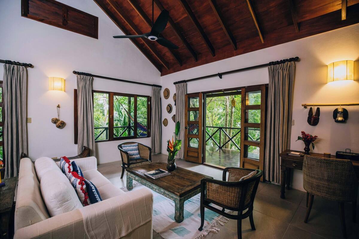 Habitación acogedora con sofá, sillas, una mesa de centro y un escritorio. Amplias ventanas con cortinas ofrecen luz natural y vistas a la vegetación exterior.
