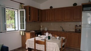 A kitchen with wooden cabinets, a dining table with chairs, a refrigerator, and a window with curtains. - Letos Villas (Paxos)