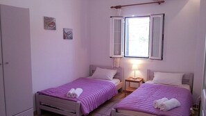 A small bedroom with two beds, each with a purple bedspread and white pillows. There is a window with open shutters, a bedside table with a lamp, and a wardrobe. - Letos Villas (Paxos)