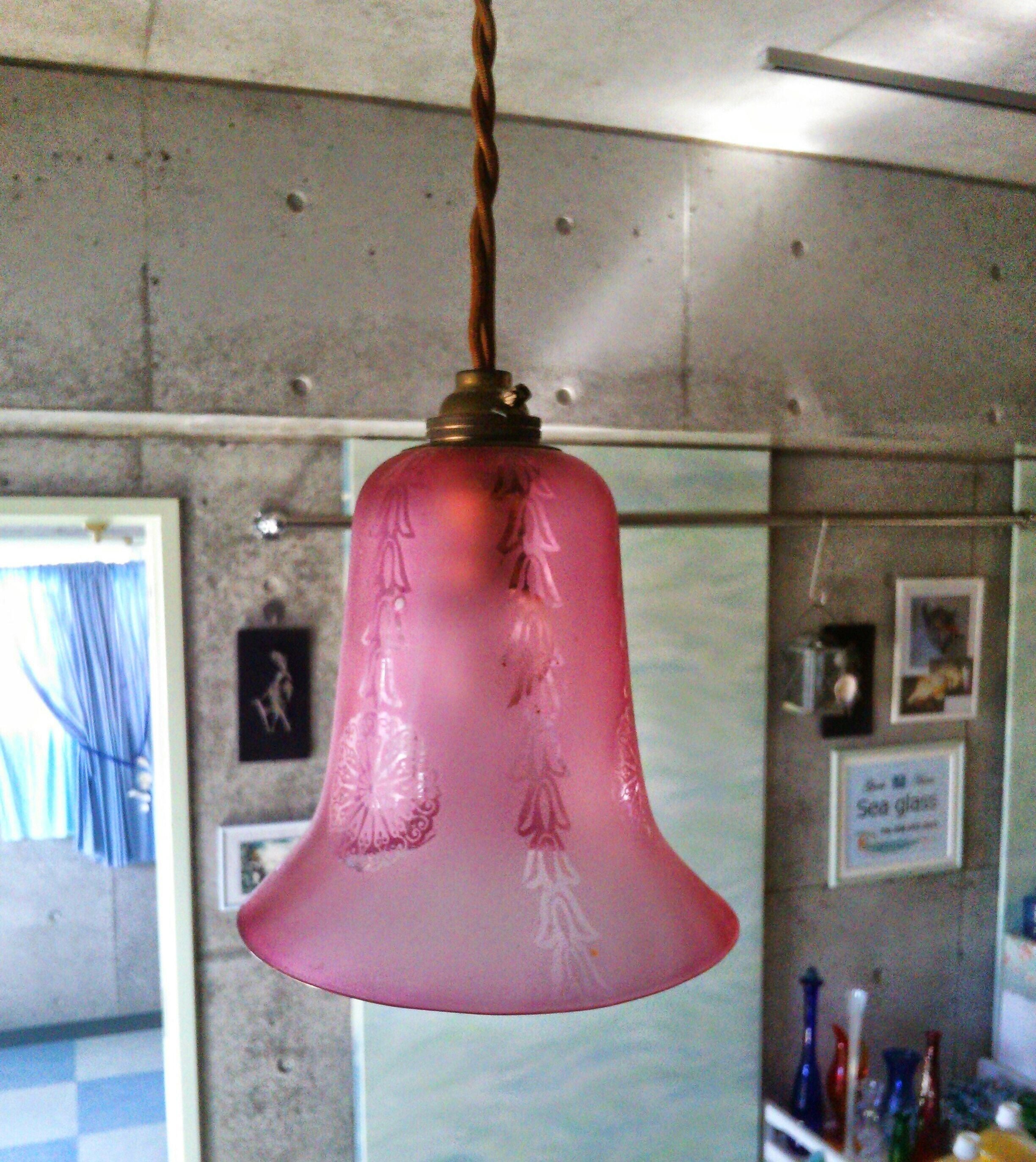 A pink bell-shaped pendant light hanging from a ceiling in a room with a concrete wall and a window with blue curtains.