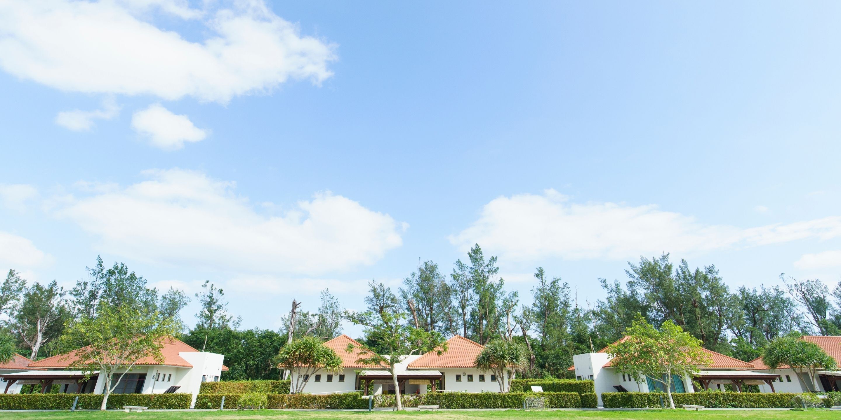 A row of white buildings with red roofs, surrounded by green hedges and trees.