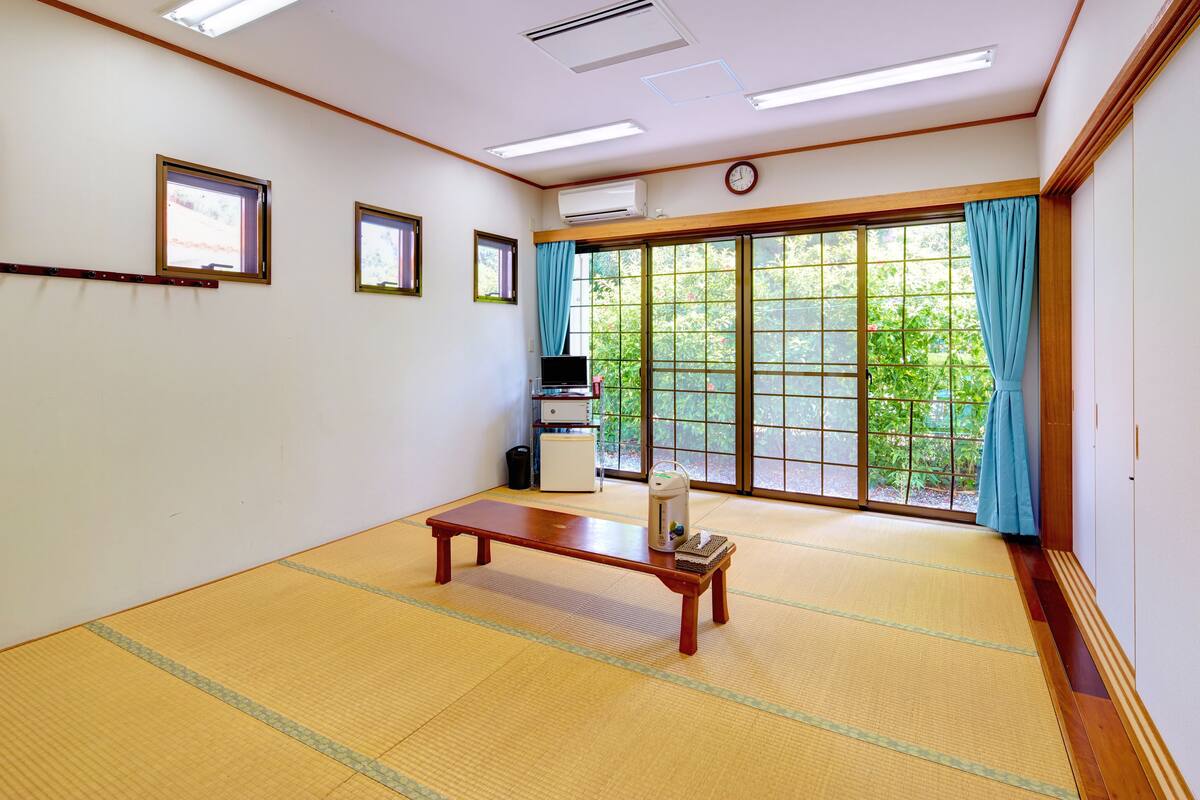 A room with tatami flooring, a wooden bench, a small table with a kettle, a heater, a window with blue curtains, and a clock on the wall.