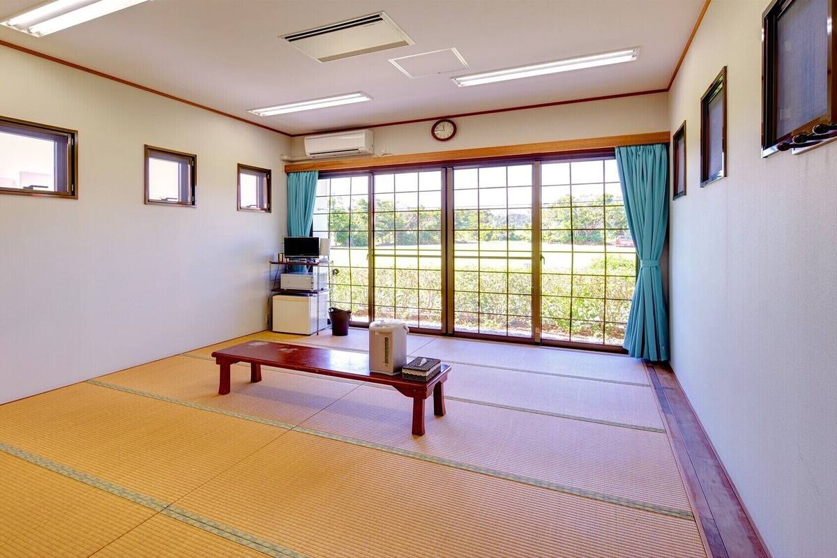 A room with tatami flooring, a wooden bench, a television on a stand, and a large window with curtains.
