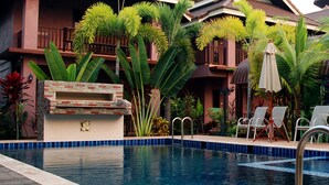 Piscina con sillas de descanso y sombrillas, rodeada de plantas tropicales y un edificio con balcones.