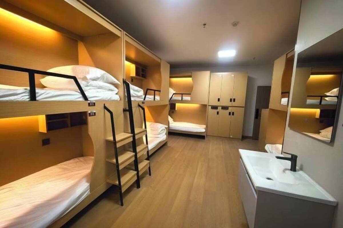 A dormitory room with bunk beds, a small desk, a mirror, and a sink.