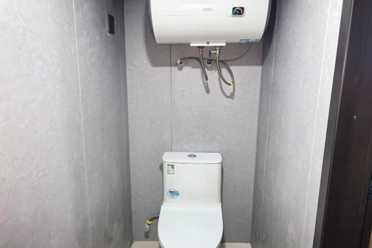 A compact, modern bathroom with a white toilet, grey tiled walls, and a visible water heater above the toilet.