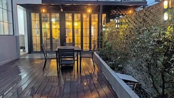 Une terrasse en bois avec une table et des chaises, un mur en briques et une porte vitrée donnant sur un espace intérieur.