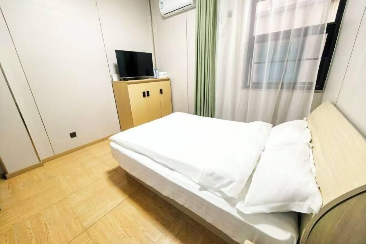 A single bed with white bedding, a television on a wooden cabinet, and a window with sheer curtains.