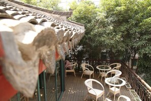Un edificio de estilo tradicional con tejado de tejas, zona de estar al aire libre con sillas y mesas de mimbre, y exuberante vegetación al fondo.