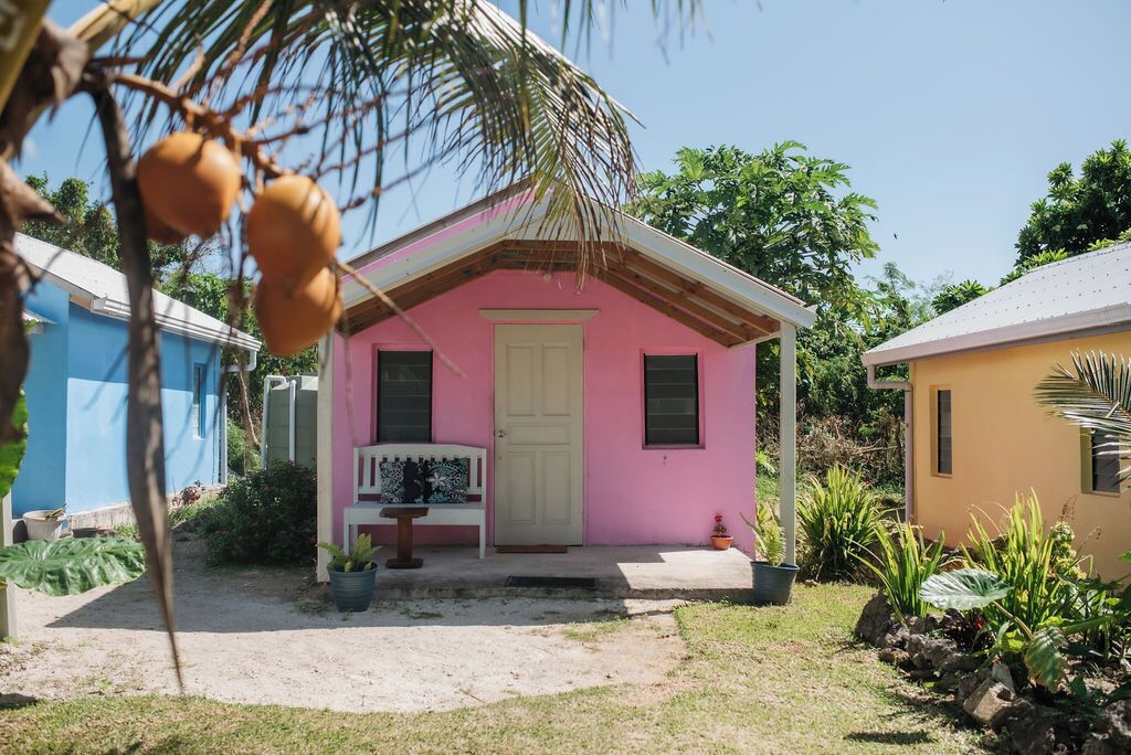 Een klein, roze huis met een witte deur, een bankje en een bloempot voor.
