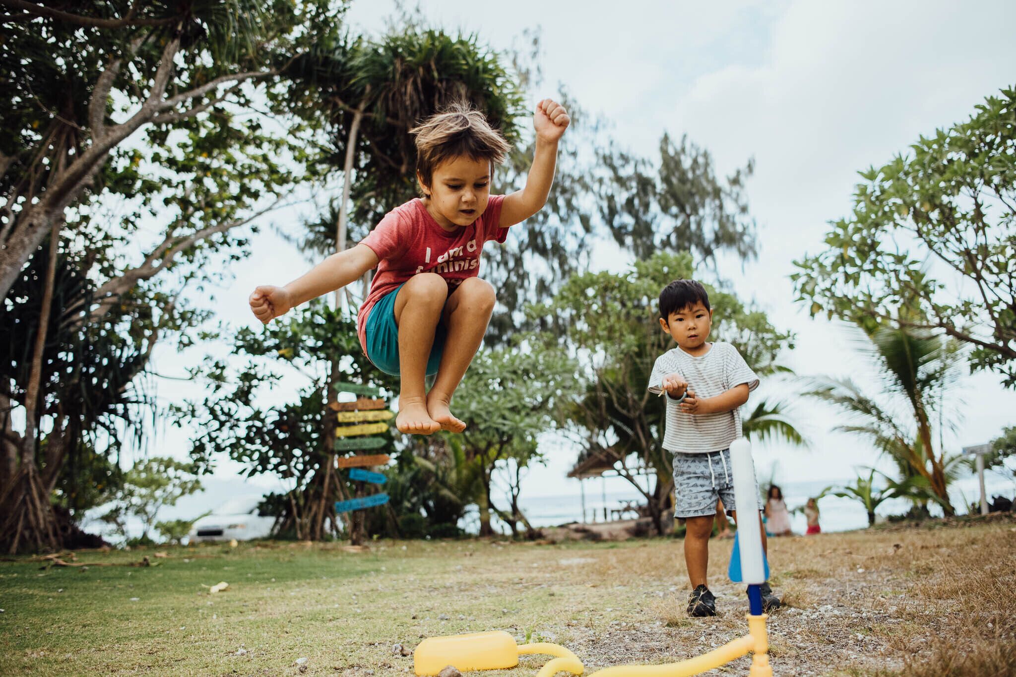 A child jumping on a trampoline in a grassy area with trees and a view of the ocean in the background.