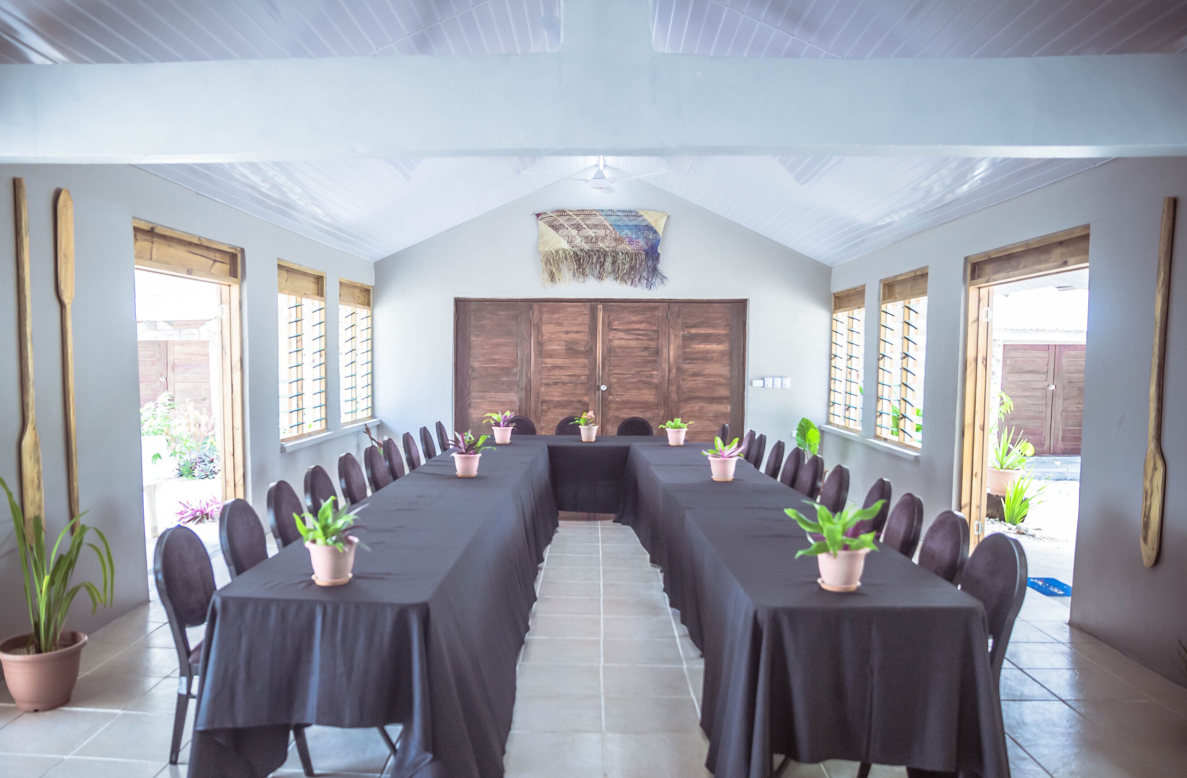 Un salón de conferencias con una larga mesa, sillas, plantas en macetas y puertas de madera.