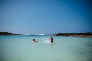 Two people playing in clear turquoise water with a rocky shoreline and a clear sky in the background. - Le Life Resort (Epule)