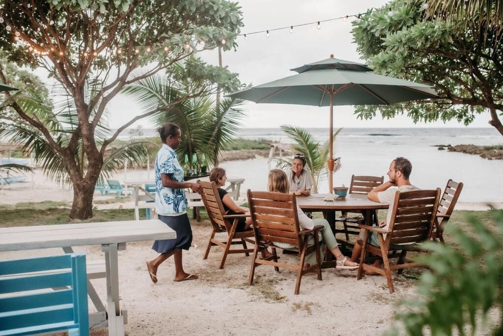 Un grupo de personas sentadas en una mesa de madera, bajo un paraguas, al aire libre con vista al mar.