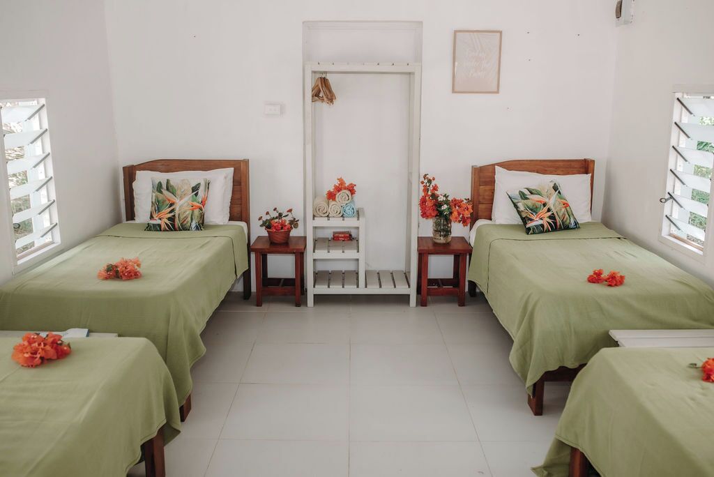 Una habitación con dos camas, cada una con una colcha verde y un cabecero de madera. Hay mesitas con adornos y un estante blanco con toallas dobladas.