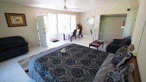 A bedroom with a large bed, a sofa, a desk, and a chair. There is a door, a window, and a ceiling fan.