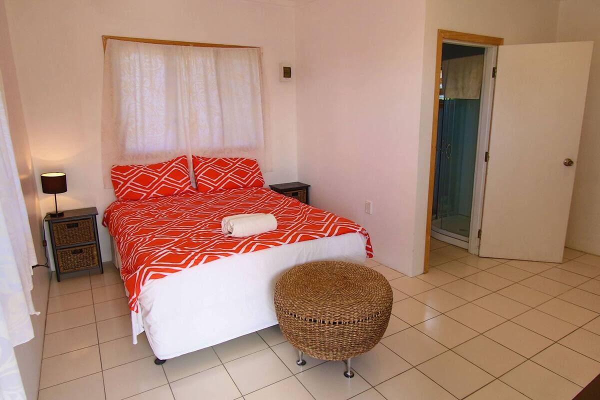 A bedroom with a bed, a bedside table, a lamp, a wicker ottoman, and a door leading to another room.