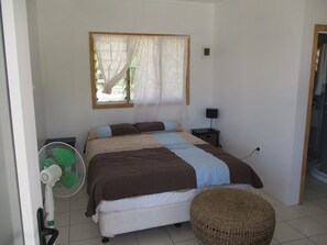 A bedroom with a bed, a bedside table with a lamp, a standing fan, and a woven stool. - White Sands Beach Resort Tonga (Kanokupolu)