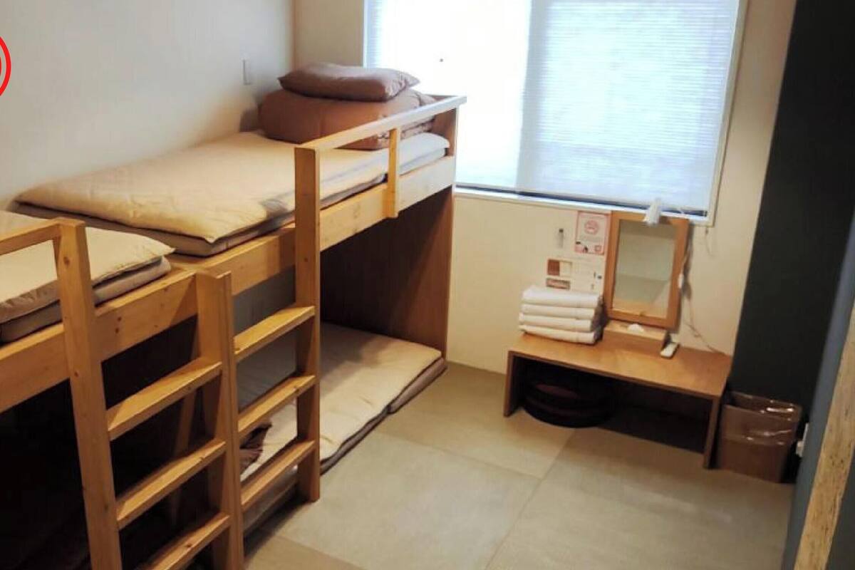 Habitación tipo dormitorio pequeña con literas, un escritorio reducido y una ventana con persianas.