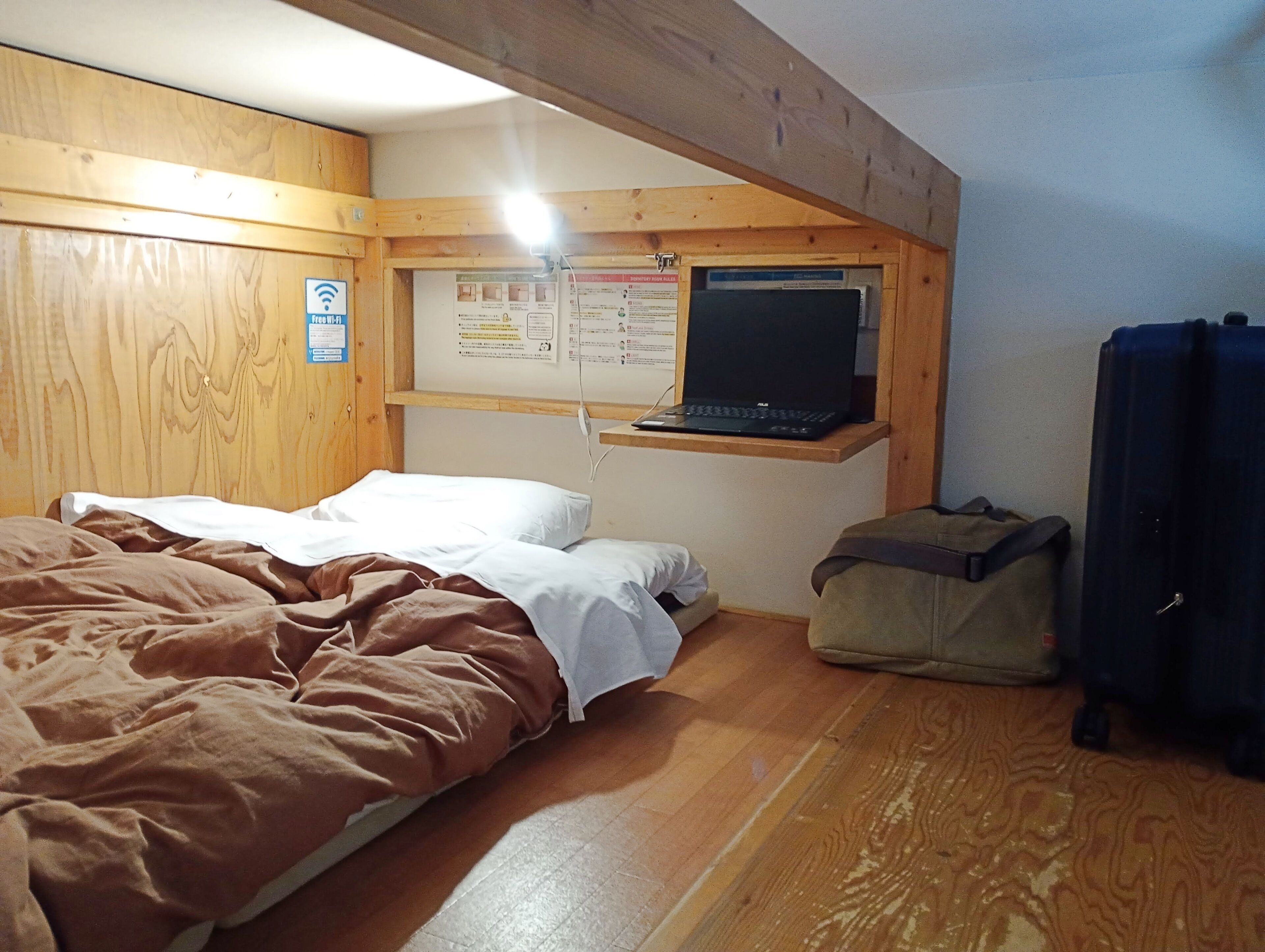 A room with a bed, a desk with a laptop, a bulletin board, and a bag.