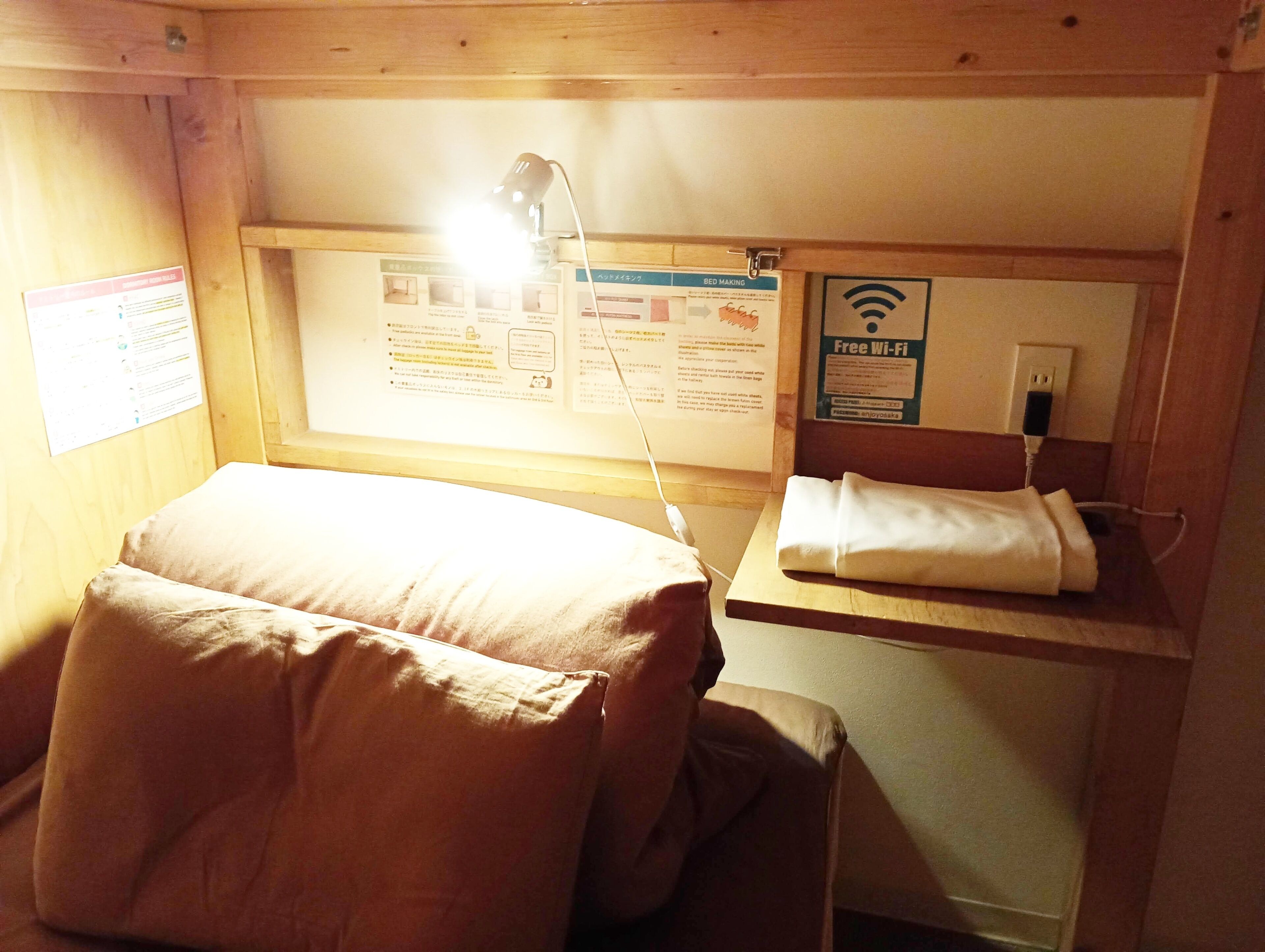 A hotel room with a bed, a desk with a lamp, a chair, a bulletin board with information, and a sign indicating free Wi-Fi.