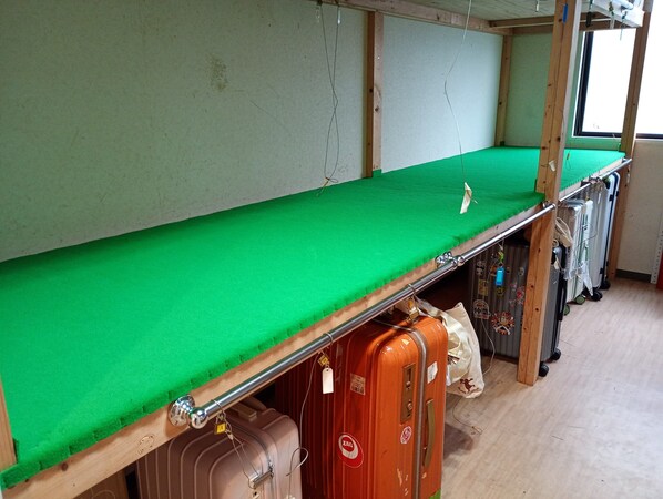 Uma cama beliche com colchão verde, estrutura de madeira e barra de metal. Há malas embaixo da cama e uma janela com cortinas.