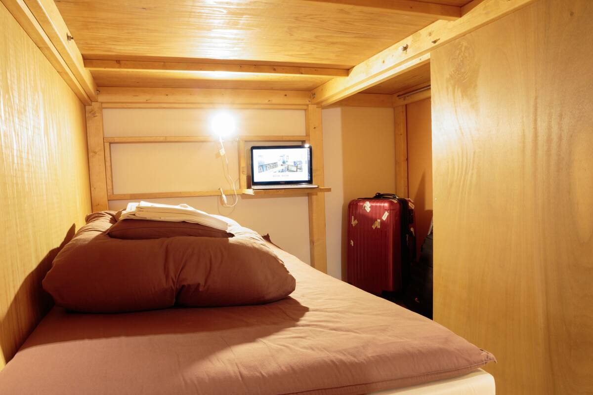 Una habitación pequeña y funcional con cama, televisor en la pared y una maleta apoyada en la pared.