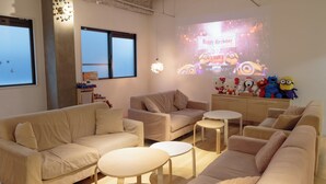 A modern living room with beige sofas, a small round table, and a projector screen displaying a birthday message. - J-Hoppers Osaka Universal - Hostel (Osaka)