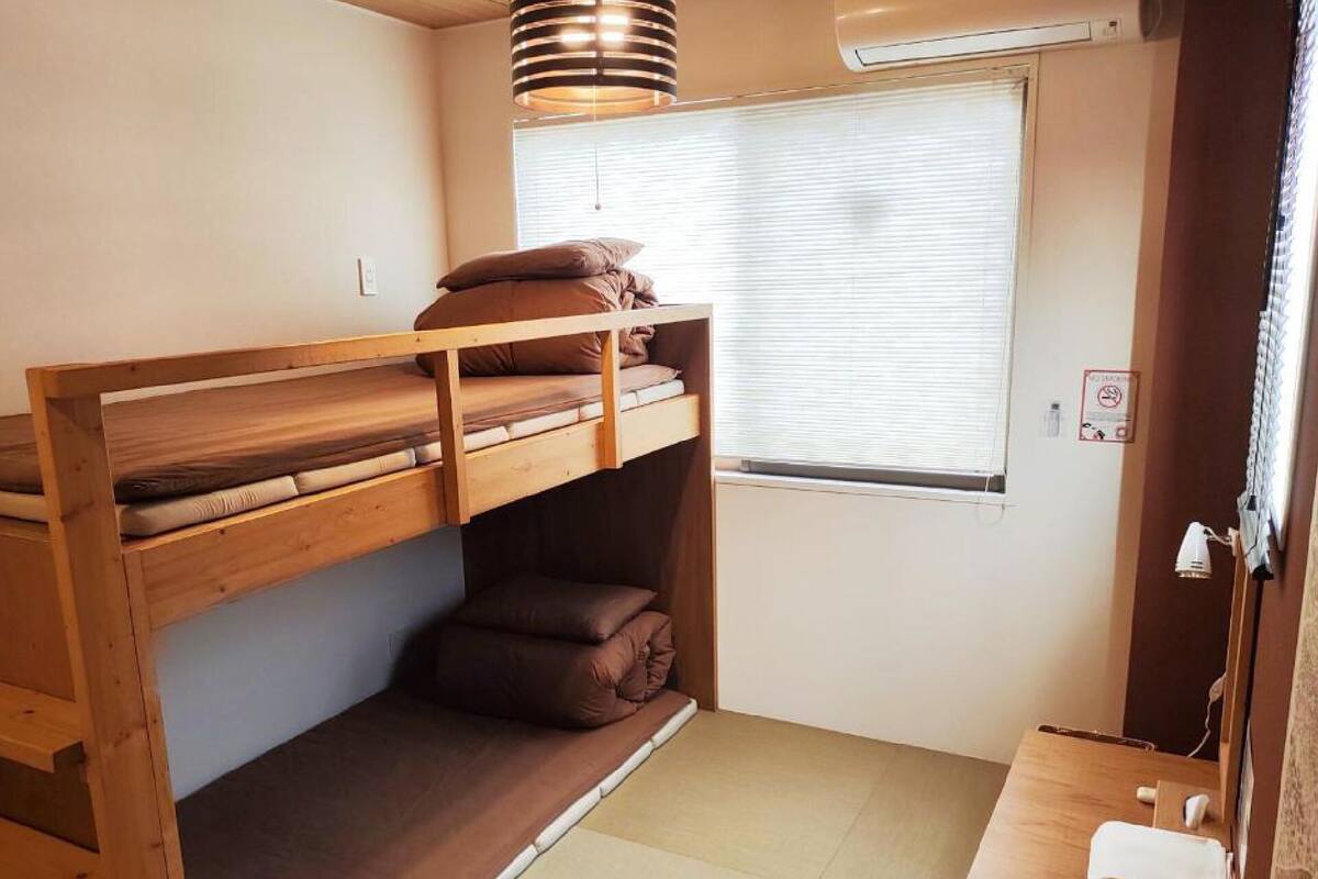 Una habitación pequeña y funcional con una cama litera, una ventana con persianas, una mesita con una toalla y una lámpara en el techo.