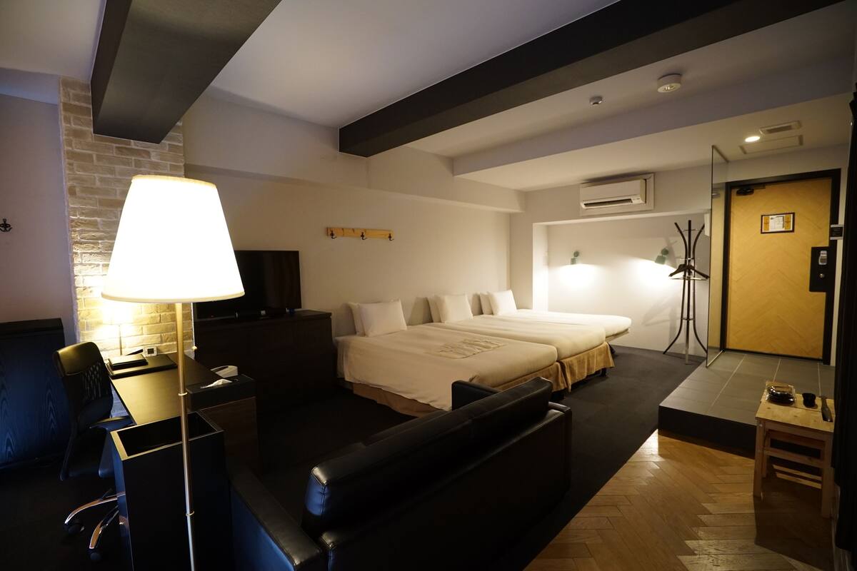 A modern hotel room with a large bed, a desk, and a sofa. There is a lamp, a TV, and a coat rack.
