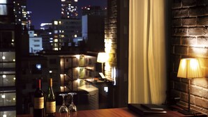 A table with two wine bottles and two wine glasses, a lamp, and a laptop in a room with a brick wall and a view of a cityscape at night.