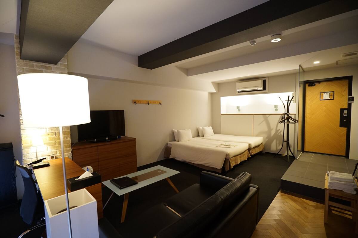 A modern hotel room with a bed, a desk with a lamp, a TV, and a wardrobe.