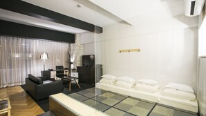 A modern hotel room with a sofa, a small desk, a bed with pillows, and a glass partition.