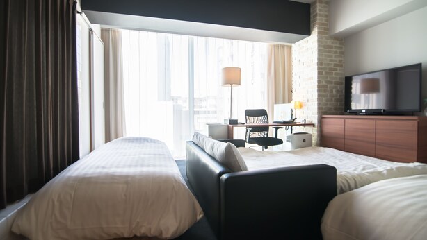 A hotel room with two beds, a desk with a chair, a television, and a large window with curtains.