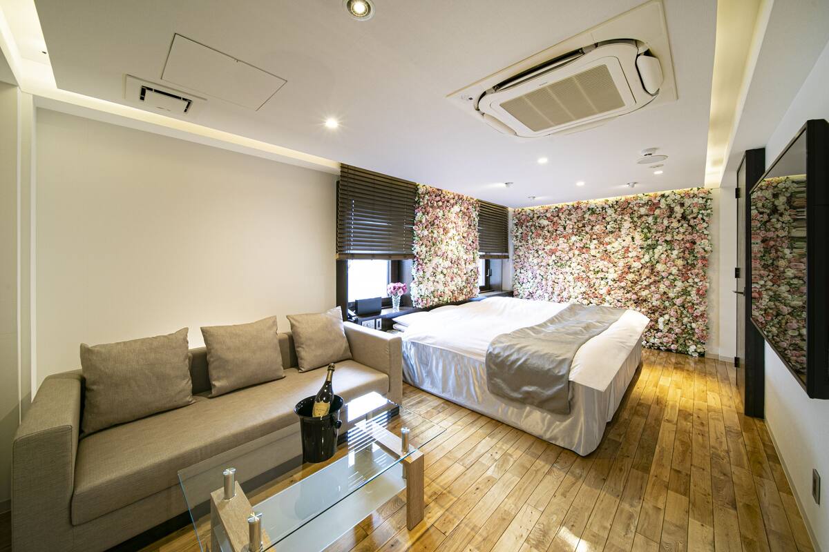 Une chambre d’hôtel moderne avec un plancher en bois, un lit, un canapé, une table basse en verre et un mur décoré d’un papier peint à motifs floraux.