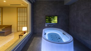 A modern bathroom with a freestanding bathtub, a television mounted on the wall, and a wooden bench with a lamp.