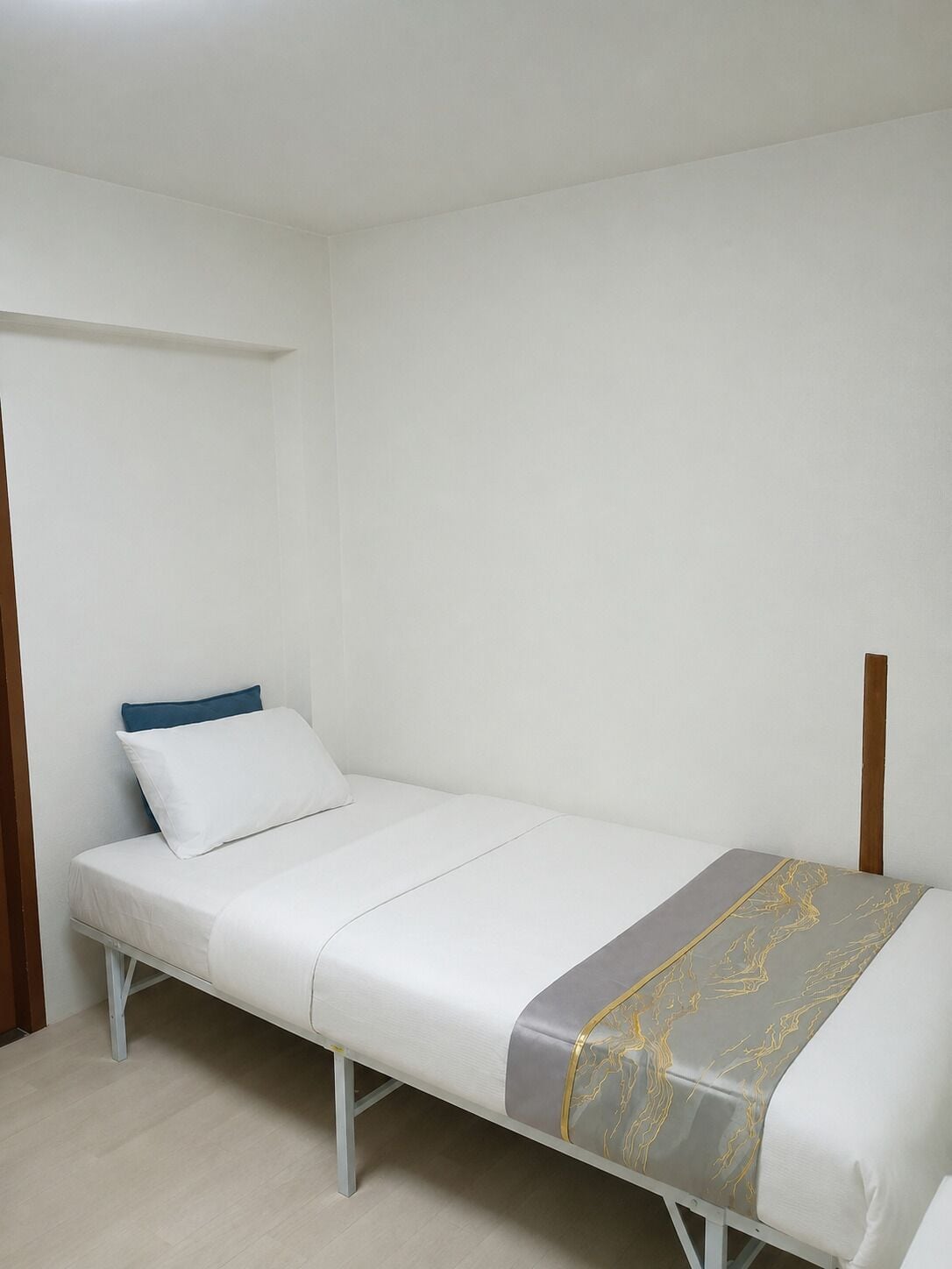A single bed with a white mattress, a blue pillow, and a grey blanket with gold patterns.