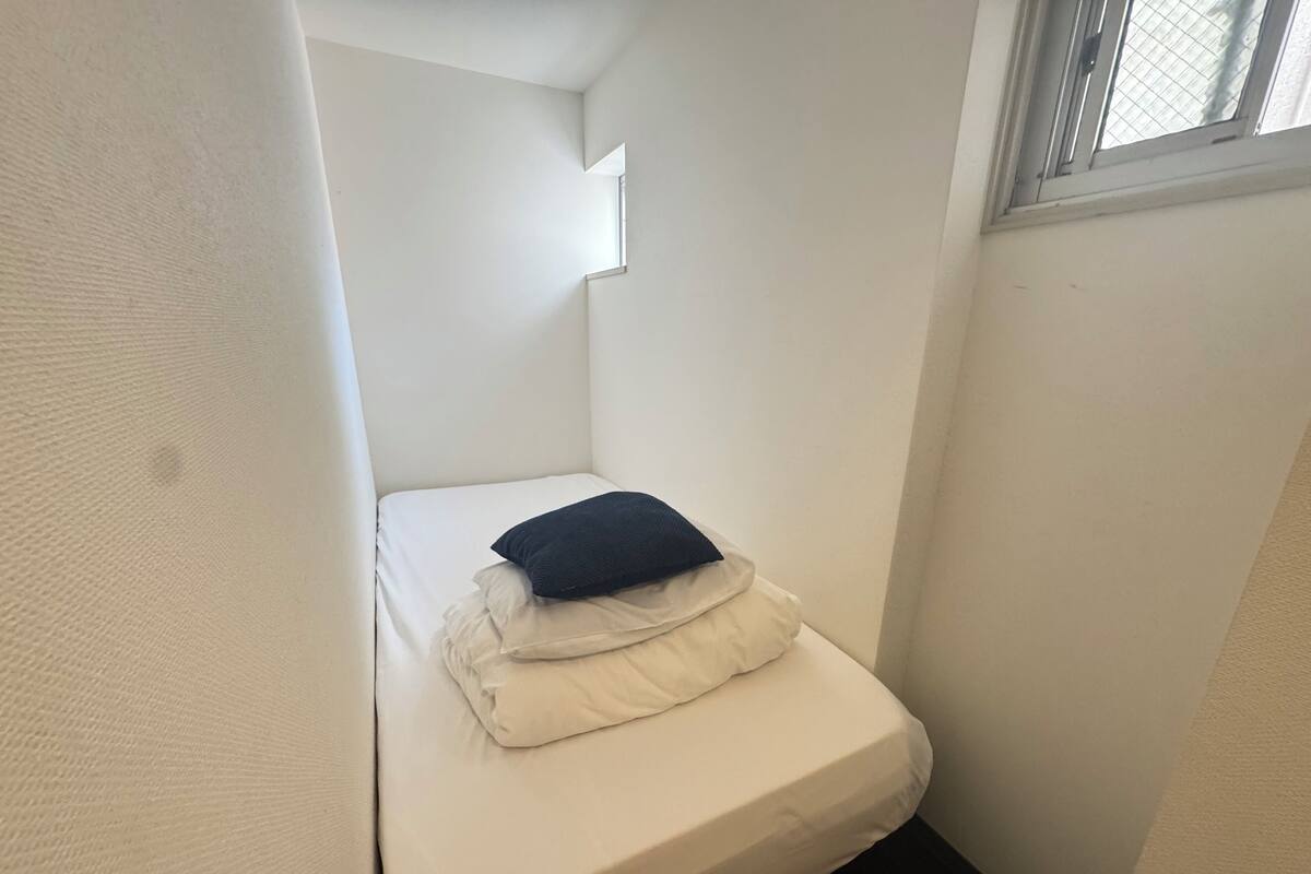 A small, narrow room with a single bed, a window, and a white wall.
