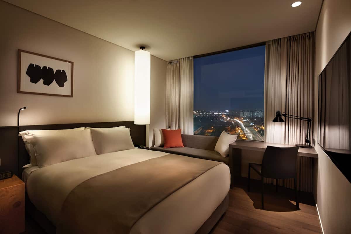 A hotel room with a large bed, a desk, and a chair. There is a window with a city view.