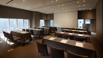 A conference room with multiple tables and chairs, large windows, and a projector screen.