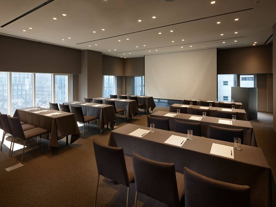 A conference room with multiple tables and chairs, large windows, and a projector screen.