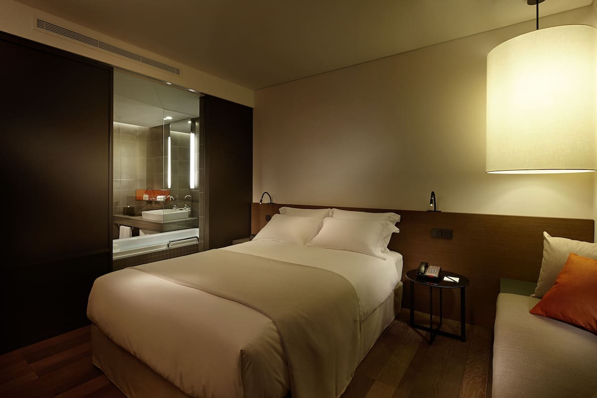 A hotel room with a large bed, a bedside table with a lamp, a small table with a phone, and a bathroom visible through a glass partition.