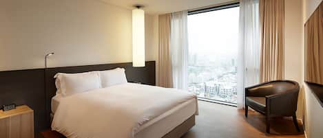 A hotel room with a large bed, a desk, a chair, a nightstand, and a view of the city through the window.
