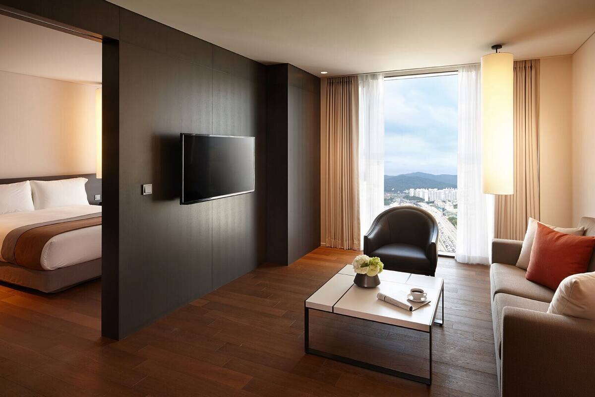 A hotel room with a bed, a TV mounted on the wall, a coffee table with a vase and a cup, a sofa, and a view of a cityscape through large windows.