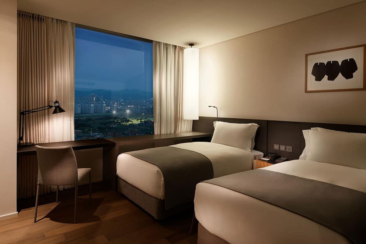 A hotel room with two beds, a desk, a chair, and a large window offering a city view.