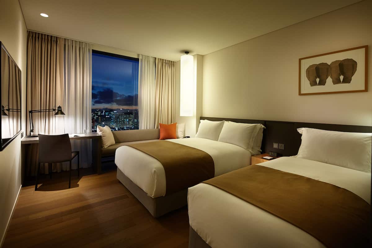 A hotel room with two beds, a desk, a chair, and a large window with a city view.